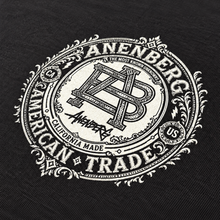 Close-up of the Anenberg Truth & Honor Black T-Shirt featuring an intricate white Anenberg American Trade crest with an ornate AB monogram, decorative Victorian-style flourishes, The Most Known Unknowns lettering, and California Made branding. Detailed vintage-inspired screen-printing on black fabric.
