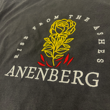 Angled close-up of the Anenberg From the Ashes Tee showcasing detailed golden yellow rose embroidery with intricately stitched petals, leaves, and stem rising above bright red flames. White embroidered Rise From the Ashes lettering curves around the rose, with the bold ANENBERG wordmark stitched beneath. Visible raised threadwork and soft vintage-washed charcoal cotton fabric highlight the premium embroidery and craftsmanship of this oversized heavyweight T-shirt from Southern California streetwear brand An