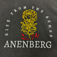 Close-up of the Anenberg From the Ashes Tee featuring detailed golden yellow rose embroidery with individually stitched petals, leaves, and stem rising above vibrant red flames. White embroidered Rise From the Ashes lettering curves around the floral design, with the bold ANENBERG wordmark stitched below. Visible embroidery thread texture and vintage-washed charcoal fabric highlight the craftsmanship of this oversized heavyweight T-shirt from Southern California streetwear brand Anenberg.