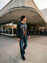 Lifestyle photo of a male model wearing the Anenberg Air Guitar Black Crystal Wash Long Sleeve T-Shirt featuring metallic-style blue and fiery orange Anenberg lettering with a lightning bolt light bulb graphic. Paired with blue jeans and black sneakers outside a modern storefront at sunset, showcasing California streetwear fashion.